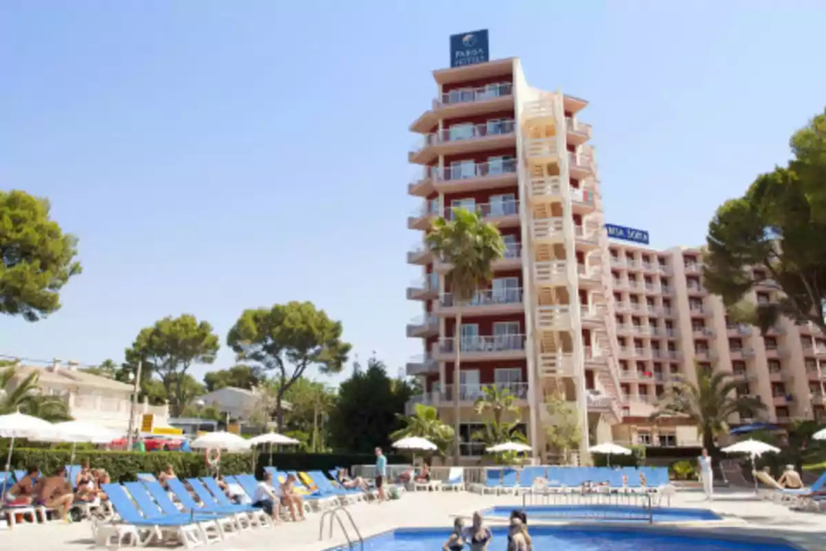 Edificio de hotel con piscina al aire libre y tumbonas azules rodeado de árboles y personas disfrutando del sol.