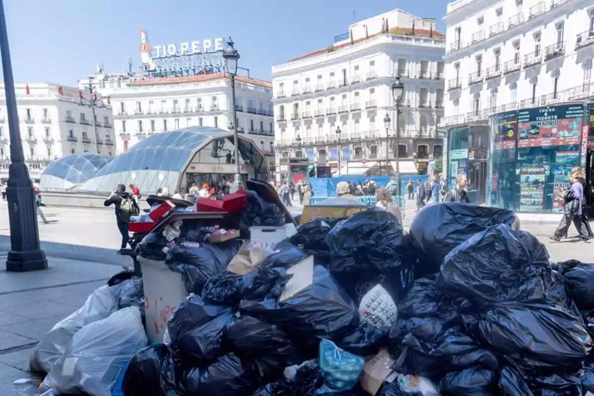 Una gran pila de bolsas de basura negras y otros desechos se acumula en una concurrida plaza urbana con edificios y un cartel visible al fondo. Una gran pila de bolsas de basura negras y otros desechos se acumula en una concurrida plaza urbana con edificios y un cartel visible al fondo.