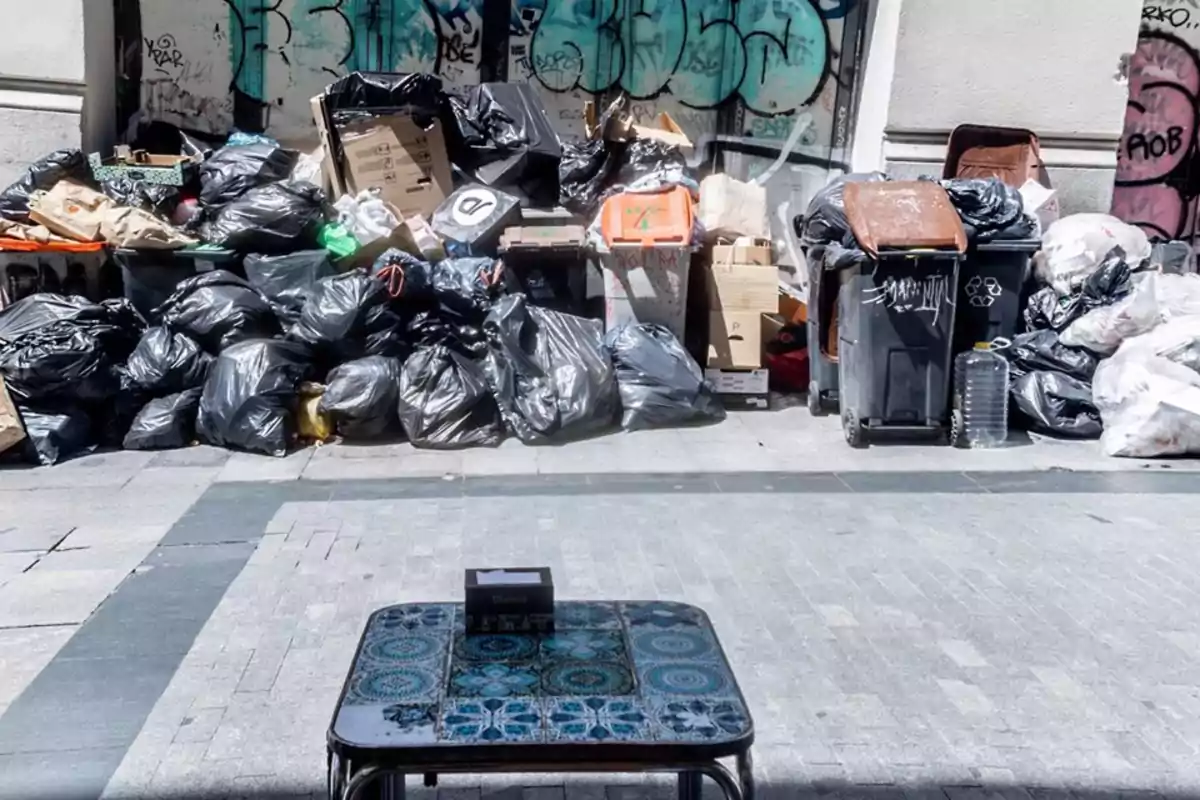 Una gran cantidad de bolsas de basura negras y cajas de cartón se acumulan en la acera frente a un muro con grafitis, mientras que en primer plano se observa una mesa pequeña con un diseño de mosaico azul. Una gran cantidad de bolsas de basura negras y cajas de cartón se acumulan en la acera frente a un muro con grafitis, mientras que en primer plano se observa una mesa pequeña con un diseño de mosaico azul.