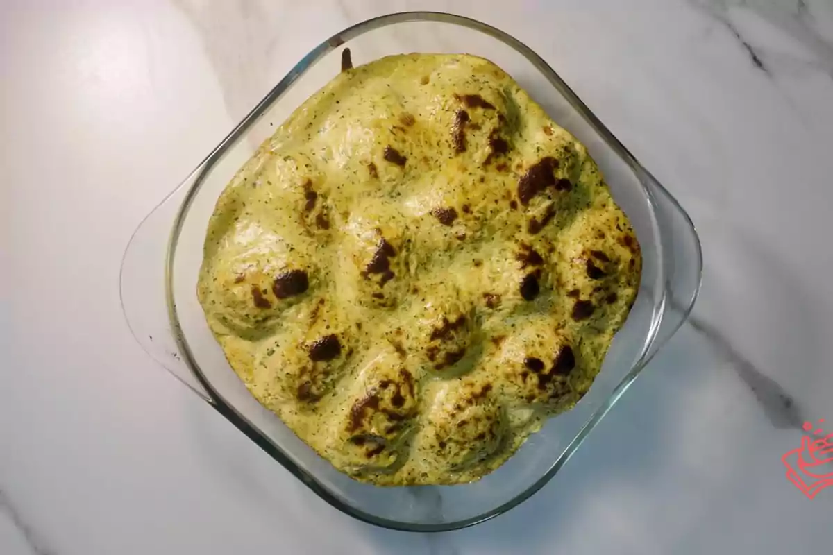 Un plato de lasaña verde con una capa superior gratinada en un recipiente de vidrio sobre una superficie de mármol blanco.