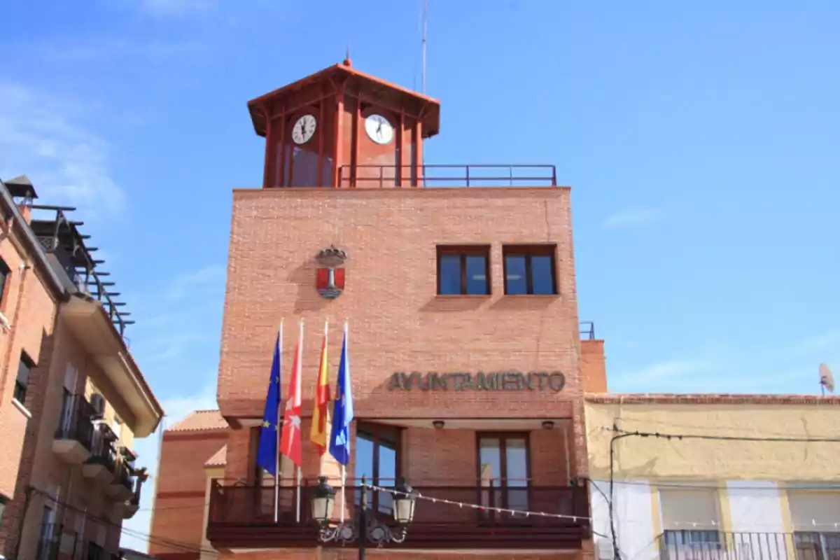 Edificio de ayuntamiento de ladrillo con torre de reloj y varias banderas en la entrada.