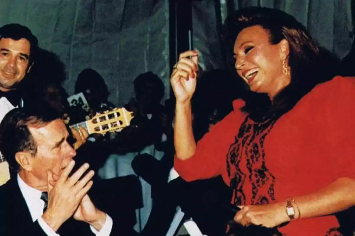 Una mujer vestida de rojo sonríe y baila mientras un hombre aplaude y otro toca la guitarra en un ambiente festivo. Una mujer vestida de rojo sonríe y baila mientras un hombre aplaude y otro toca la guitarra en un ambiente festivo.