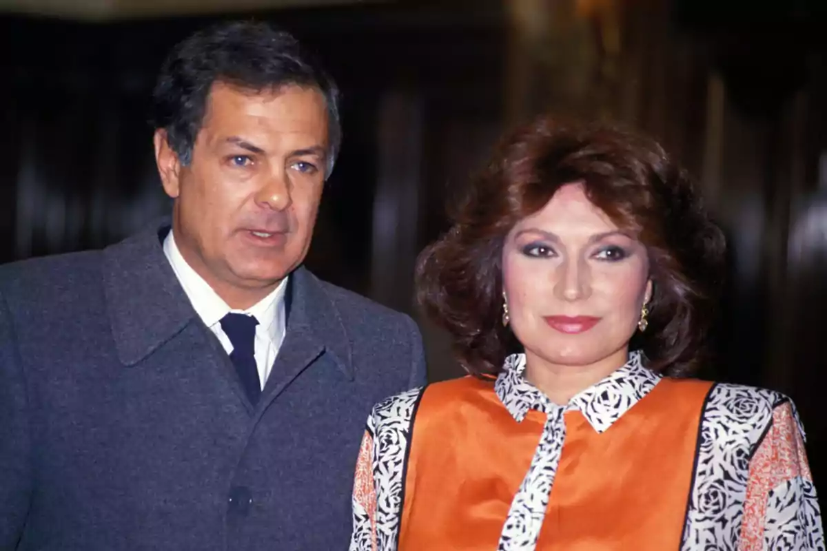 Pareja posando juntos en un evento formal ella lleva un vestido naranja con detalles en blanco y negro y él viste un abrigo gris con corbata Pareja posando juntos en un evento formal ella lleva un vestido naranja con detalles en blanco y negro y él viste un abrigo gris con corbata