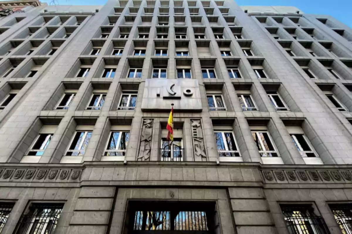 Fachada de un edificio institucional de varios pisos con una bandera de España en la entrada y ventanas simétricas