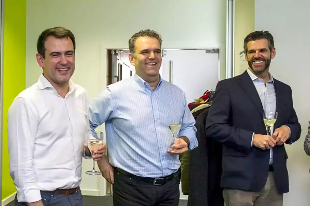 Tres hombres sonrientes sostienen copas de vino en un ambiente de oficina.