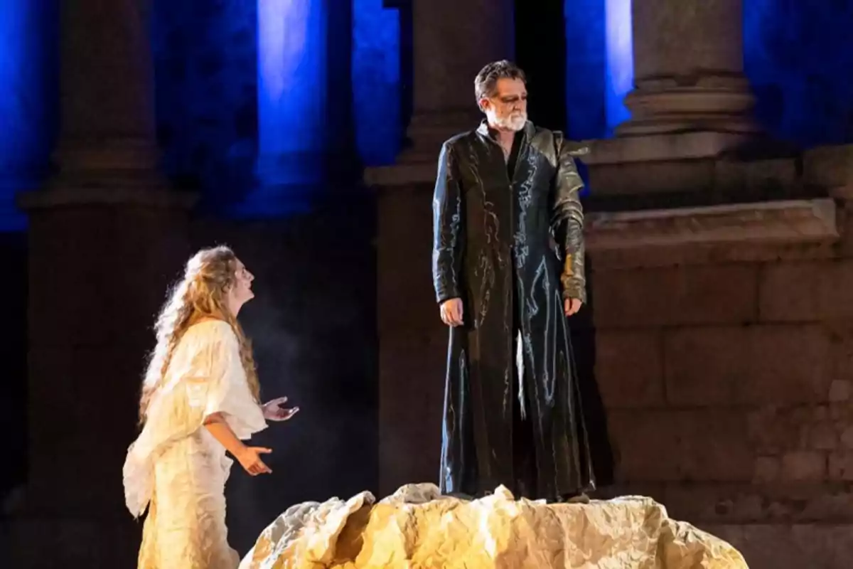 Dos actores en el escenario durante una obra de teatro, uno de pie sobre una roca con vestimenta oscura y la otra con un vestido claro mirándolo y gesticulando, con columnas iluminadas de azul al fondo Dos actores en el escenario durante una obra de teatro, uno de pie sobre una roca con vestimenta oscura y la otra con un vestido claro mirándolo y gesticulando, con columnas iluminadas de azul al fondo
