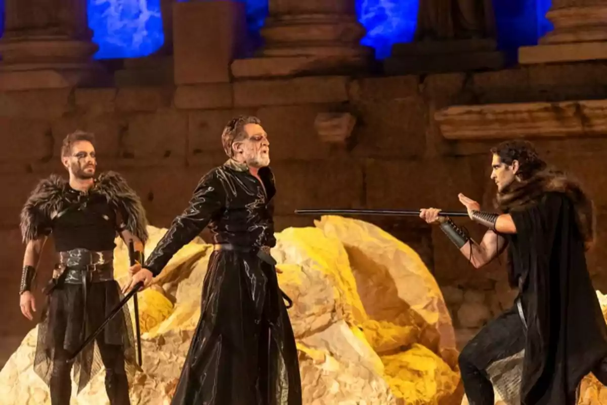Tres actores con vestuarios oscuros y capas de piel representan una escena dramática en un escenario con rocas y columnas iluminadas en tonos dorados y azules Tres actores con vestuarios oscuros y capas de piel representan una escena dramática en un escenario con rocas y columnas iluminadas en tonos dorados y azules