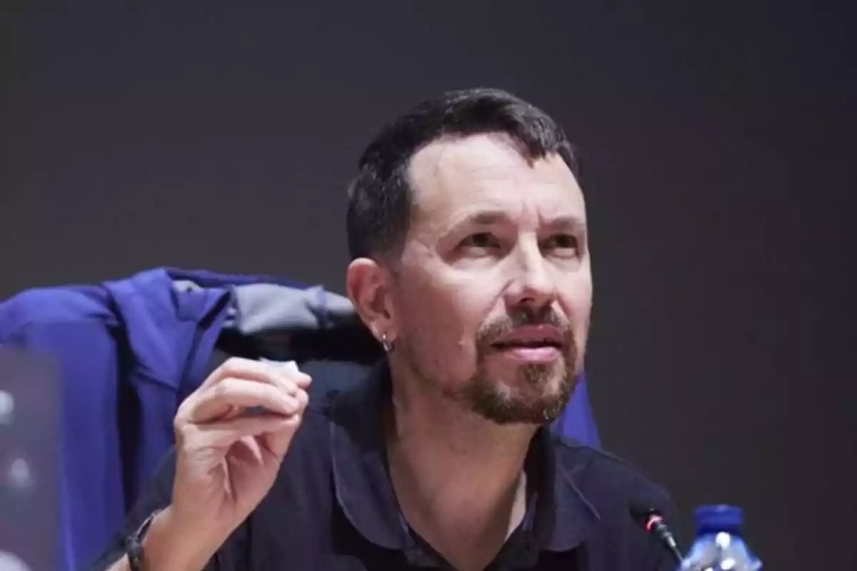 Un hombre con barba y cabello corto está hablando en un micrófono mientras gesticula con la mano. Un hombre con barba y cabello corto está hablando en un micrófono mientras gesticula con la mano.