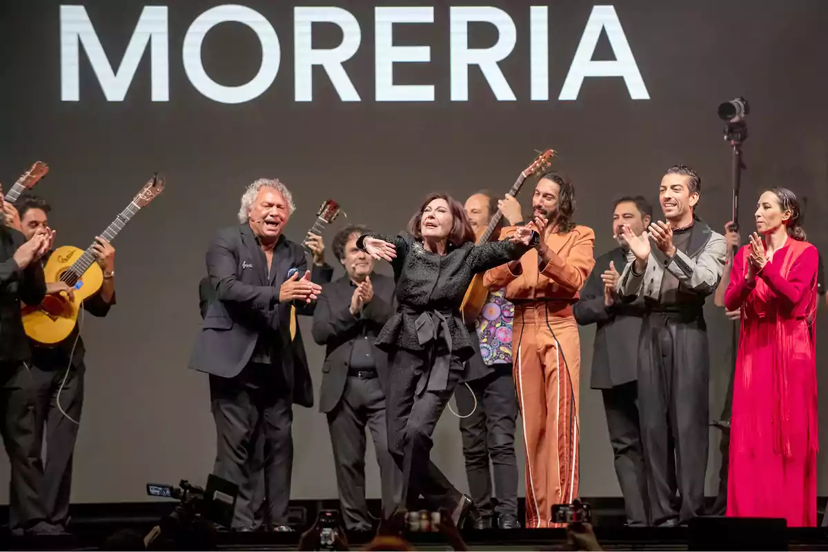 Un grupo de personas aplaude a una mujer que está bailando en un escenario con guitarristas al fondo y la palabra "MORERIA" en grande.