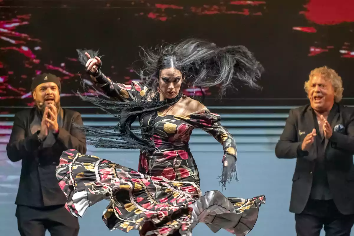 Una bailaora de flamenco con un vestido colorido realiza un movimiento enérgico mientras dos músicos aplauden en el fondo.