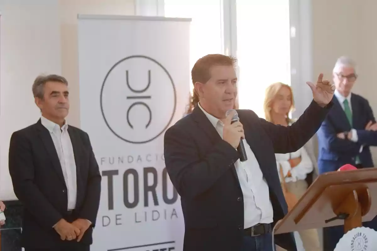 Un hombre hablando con un micrófono en un evento de la Fundación Toro de Lidia, con varias personas de pie en el fondo.