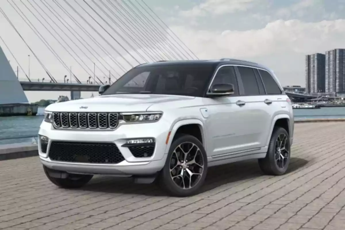 Un Jeep Grand Cherokee 4xe blanco estacionado en un área pavimentada con un puente y edificios al fondo.