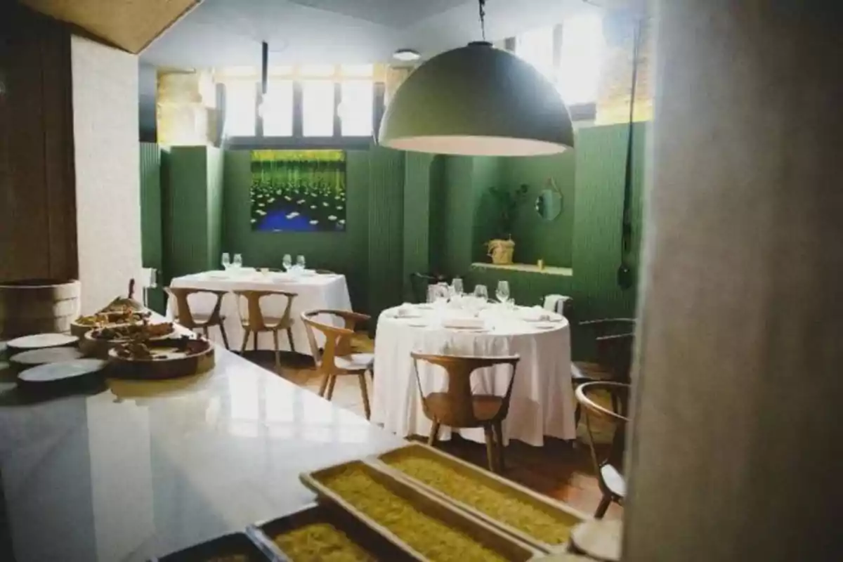 Un elegante restaurante con mesas redondas cubiertas con manteles blancos, sillas de madera y una decoración en tonos verdes, con una gran lámpara colgante y una pintura de nenúfares en la pared.