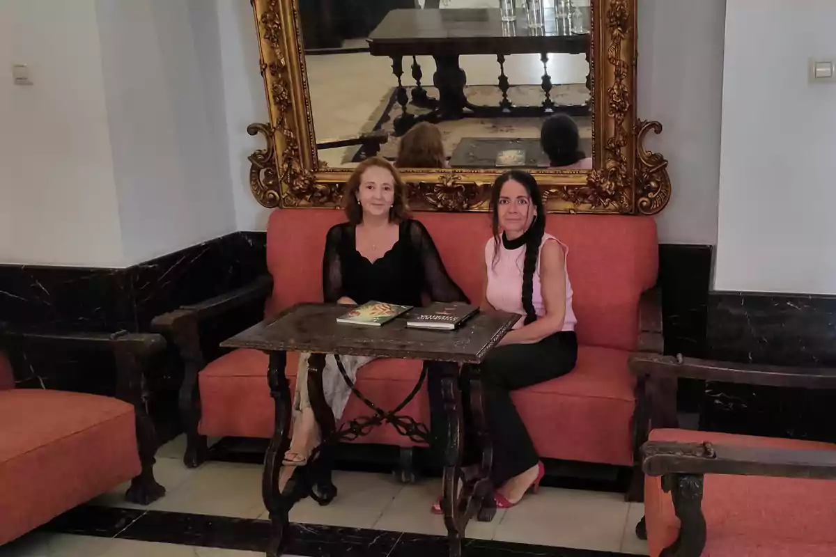 Dos mujeres sentadas en un sofá rojo frente a una mesa con libros, con un gran espejo dorado detrás.