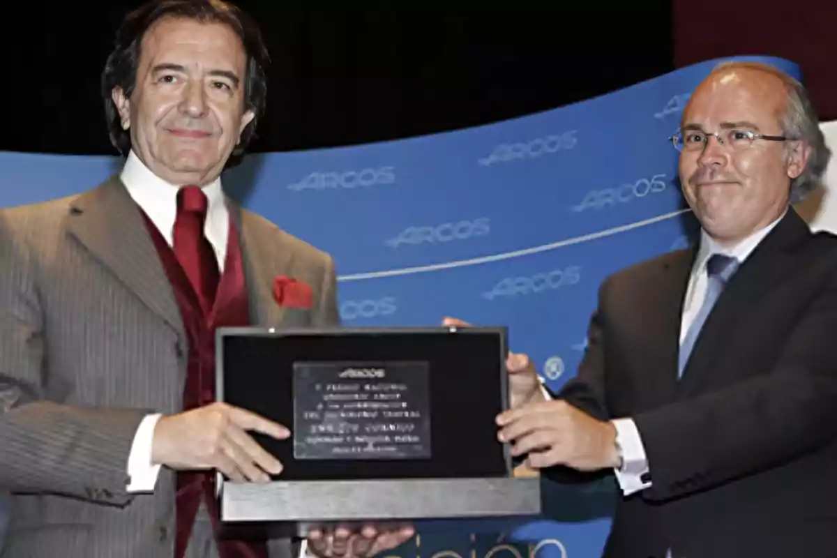 Dos hombres en traje sostienen un premio en un evento con fondo azul.