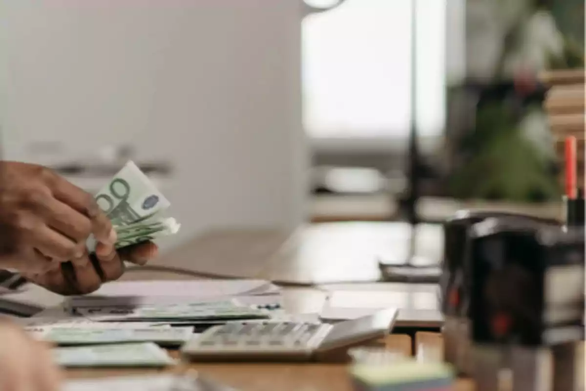 Persona contando billetes de euro en una oficina con calculadora y papeles sobre la mesa.