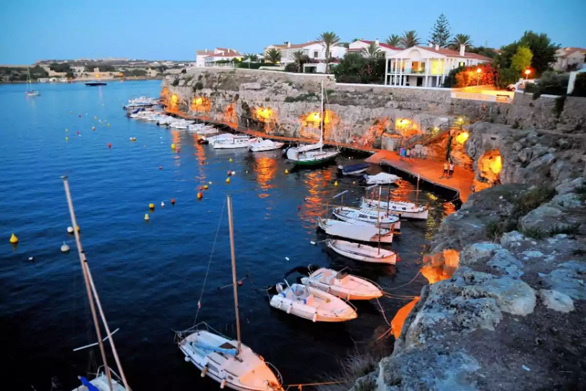 Pequeño puerto con varios barcos atracados junto a un acantilado iluminado y casas rodeadas de palmeras al fondo Pequeño puerto con varios barcos atracados junto a un acantilado iluminado y casas rodeadas de palmeras al fondo