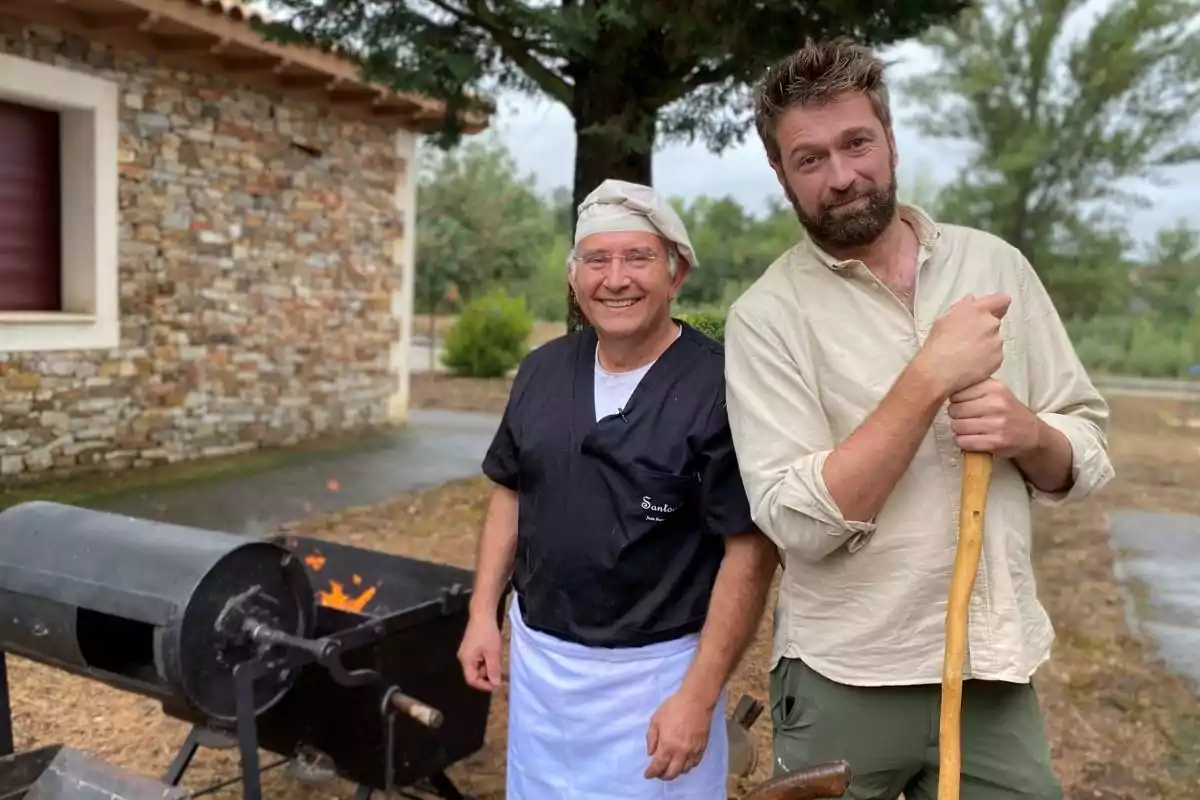 Dos hombres posan sonrientes al aire libre junto a una parrilla encendida, uno de ellos lleva ropa de chef y el otro sostiene un palo de madera.