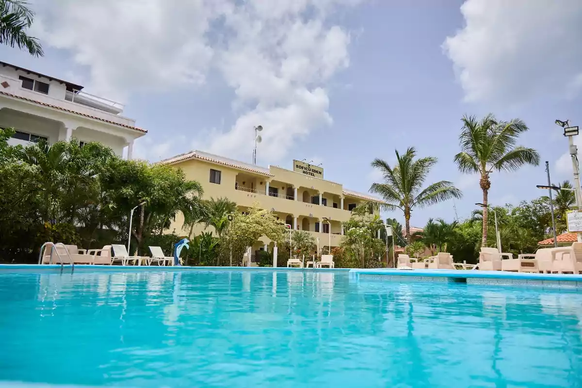 Vista de una piscina con un hotel de fondo rodeado de palmeras y vegetación bajo un cielo parcialmente nublado.