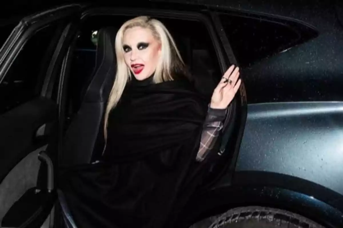 Alaska con maquillaje oscuro y cabello rubio platino está sentada en un auto, vistiendo una capa negra y saludando con la mano. Alaska con maquillaje oscuro y cabello rubio platino está sentada en un auto, vistiendo una capa negra y saludando con la mano.