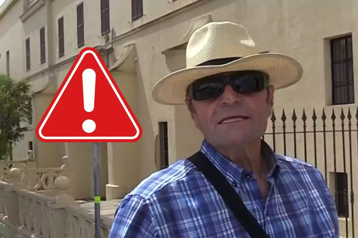 Amador Mohedano con sombrero y gafas de sol frente a un edificio beige junto a una señal de advertencia roja con un signo de exclamación