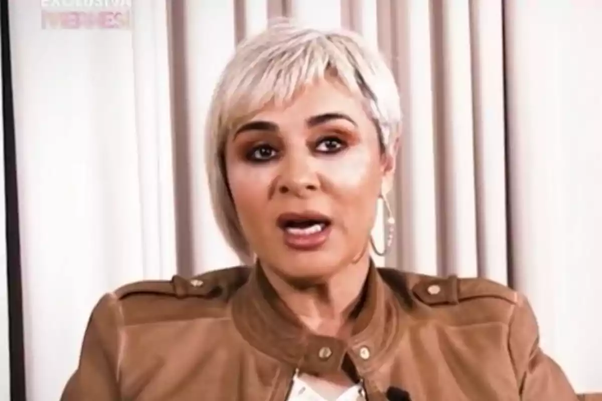 Ana María Aldón con cabello corto y rubio lleva una chaqueta marrón y está hablando frente a un fondo de cortinas claras. Ana María Aldón con cabello corto y rubio lleva una chaqueta marrón y está hablando frente a un fondo de cortinas claras.
