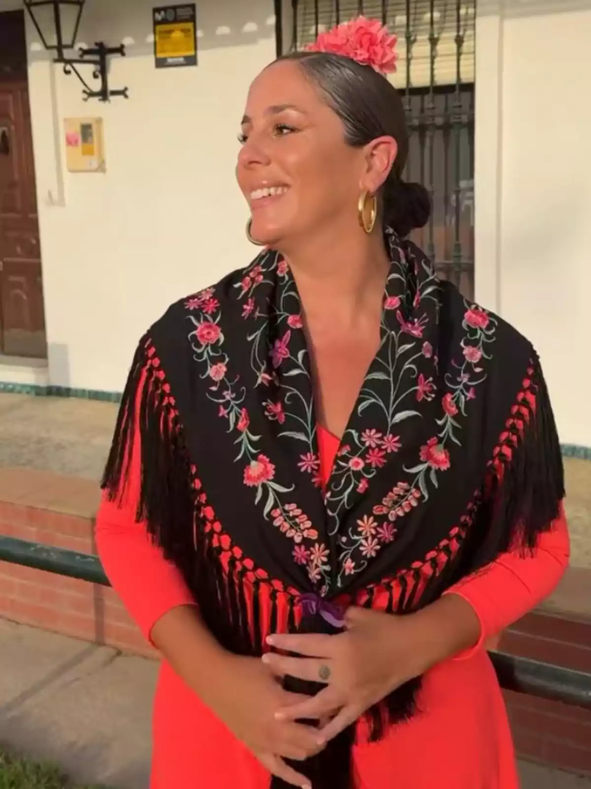 Anabel Pantoja sonriente vestida con traje tradicional flamenco de color rojo y mantón negro bordado con flores, lleva una flor rosa en el cabello y pendientes dorados, posando al aire libre frente a una casa. Anabel Pantoja sonriente vestida con traje tradicional flamenco de color rojo y mantón negro bordado con flores, lleva una flor rosa en el cabello y pendientes dorados, posando al aire libre frente a una casa.
