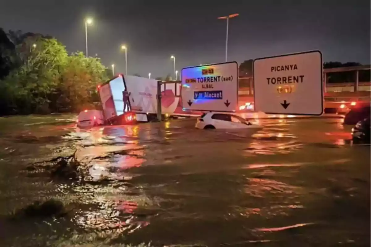 Un camión y un coche atrapados en una inundación nocturna en una carretera con señales de tráfico visibles.