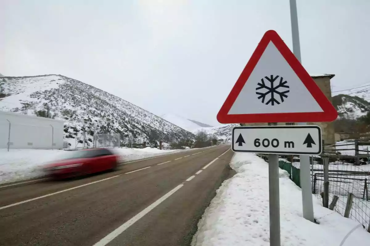 Un coche rojo pasa rápidamente por una carretera nevada con una señal de advertencia de nieve a 600 metros. Un coche rojo pasa rápidamente por una carretera nevada con una señal de advertencia de nieve a 600 metros.