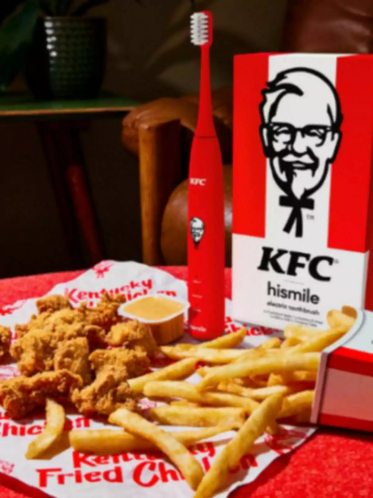 Un cepillo de dientes eléctrico rojo de KFC junto a una caja con el logotipo de la marca, acompañado de una porción de pollo frito y papas fritas sobre una servilleta con el nombre de Kentucky Fried Chicken. Un cepillo de dientes eléctrico rojo de KFC junto a una caja con el logotipo de la marca, acompañado de una porción de pollo frito y papas fritas sobre una servilleta con el nombre de Kentucky Fried Chicken.