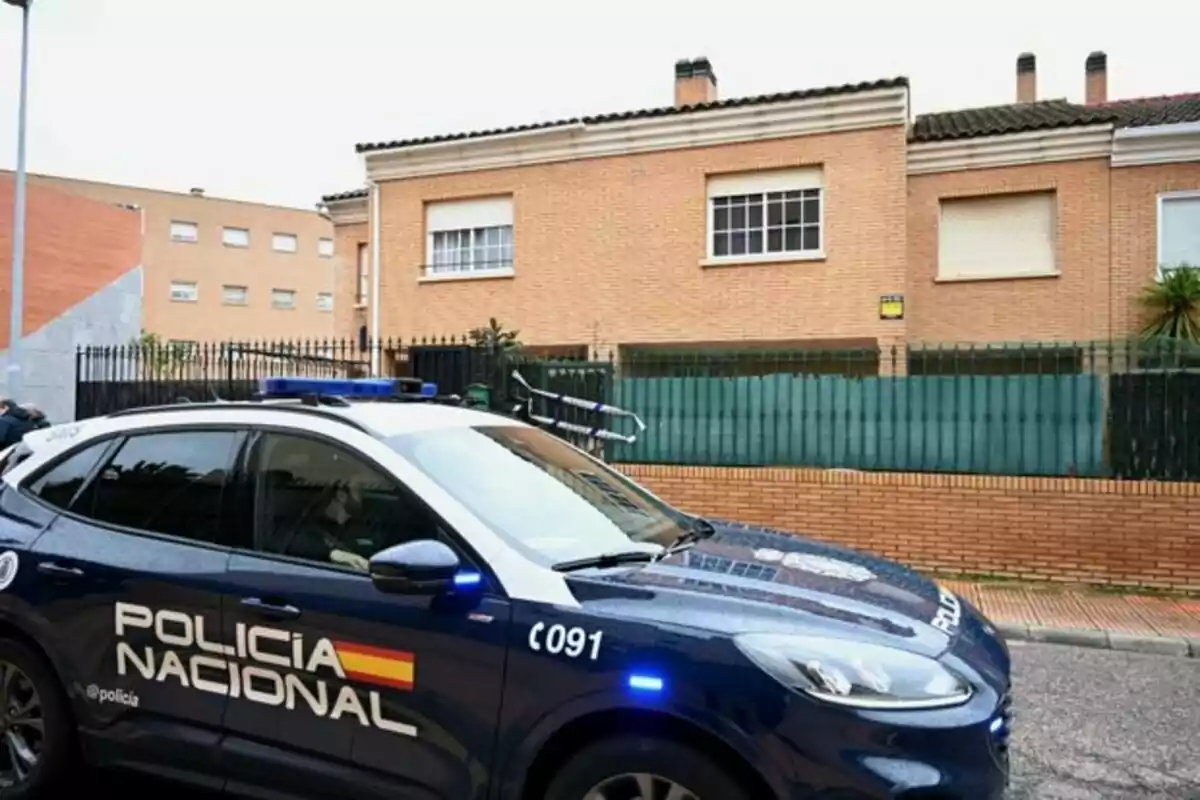 Un coche de la Policía Nacional estacionado frente a una casa de ladrillo con ventanas cerradas y una valla verde.