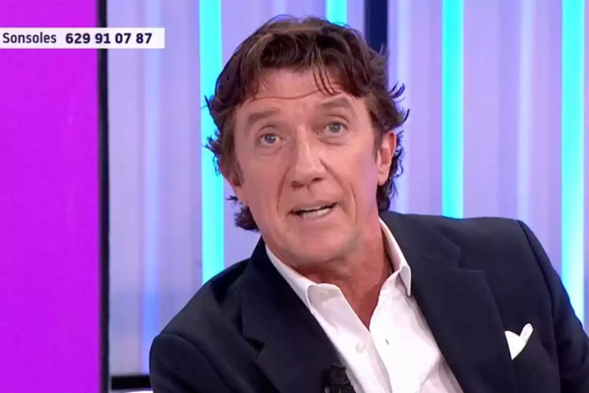 Colate de cabello castaño y ojos claros con chaqueta oscura y camisa blanca en un set de televisión con fondo de luces azules y moradas Colate de cabello castaño y ojos claros con chaqueta oscura y camisa blanca en un set de televisión con fondo de luces azules y moradas