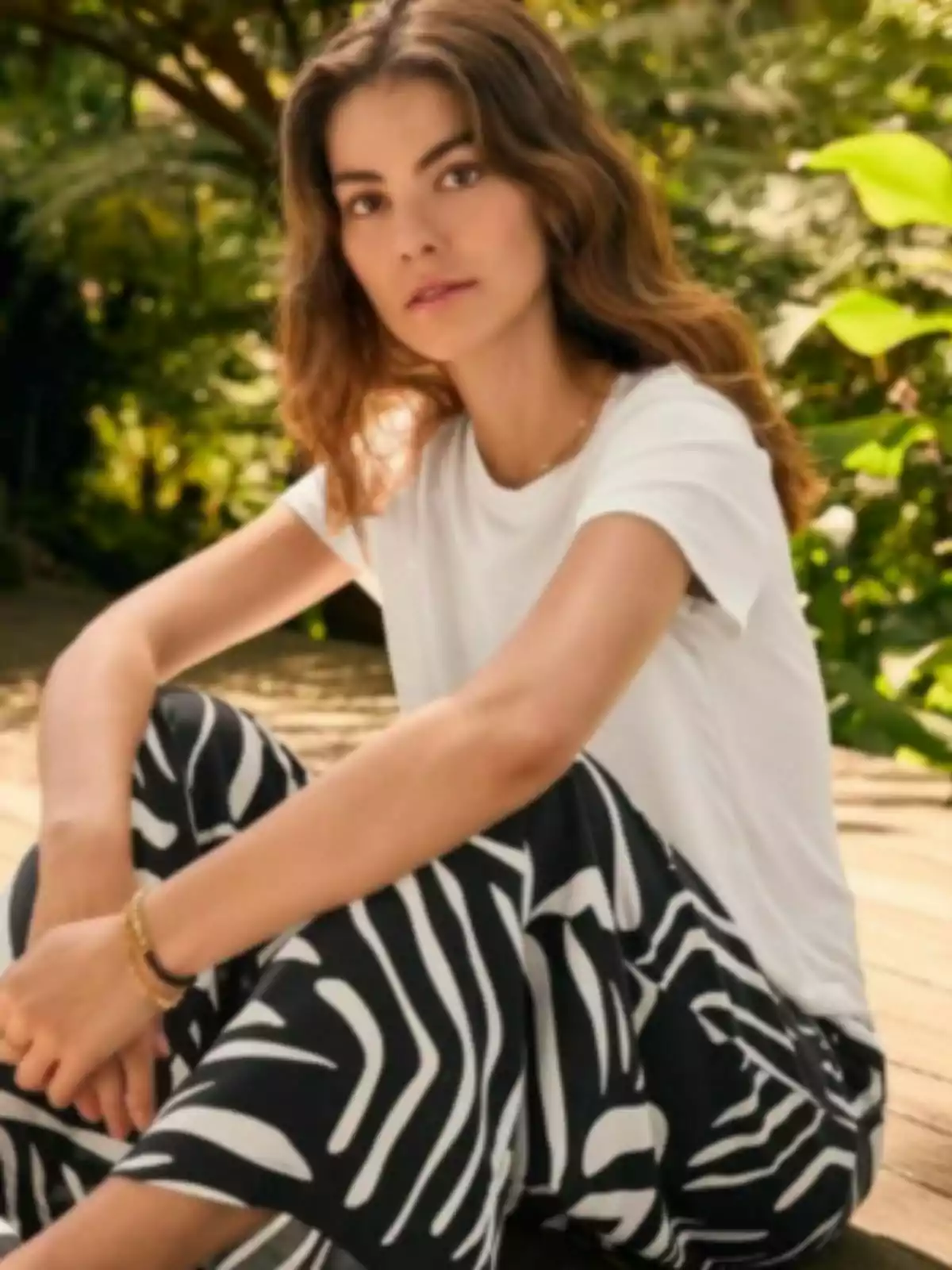 Una persona con cabello castaño y ondulado, vestida con una camiseta blanca y pantalones con estampado de rayas de Lidl, está sentada al aire libre en un entorno natural. Una persona con cabello castaño y ondulado, vestida con una camiseta blanca y pantalones con estampado de rayas de Lidl, está sentada al aire libre en un entorno natural.