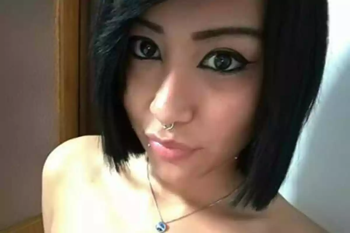 Una persona con cabello oscuro y corto, maquillaje de ojos llamativo y un piercing en la nariz, lleva un collar con un dije azul.