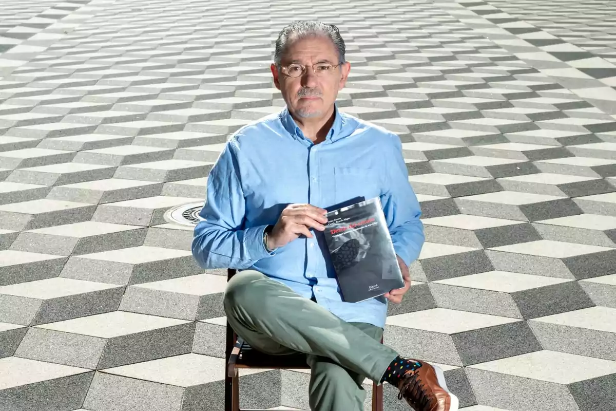 Hombre sentado en una silla sosteniendo un libro con un patrón geométrico en el suelo.