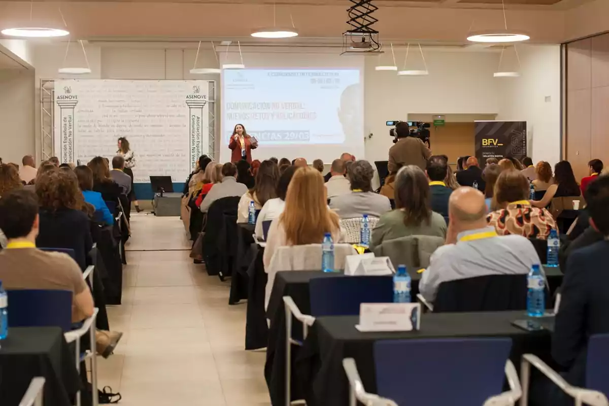 Un grupo de personas asiste a una conferencia sobre comunicación no verbal en un salón, donde una presentadora habla frente a una pantalla mientras un camarógrafo graba el evento. Un grupo de personas asiste a una conferencia sobre comunicación no verbal en un salón, donde una presentadora habla frente a una pantalla mientras un camarógrafo graba el evento.
