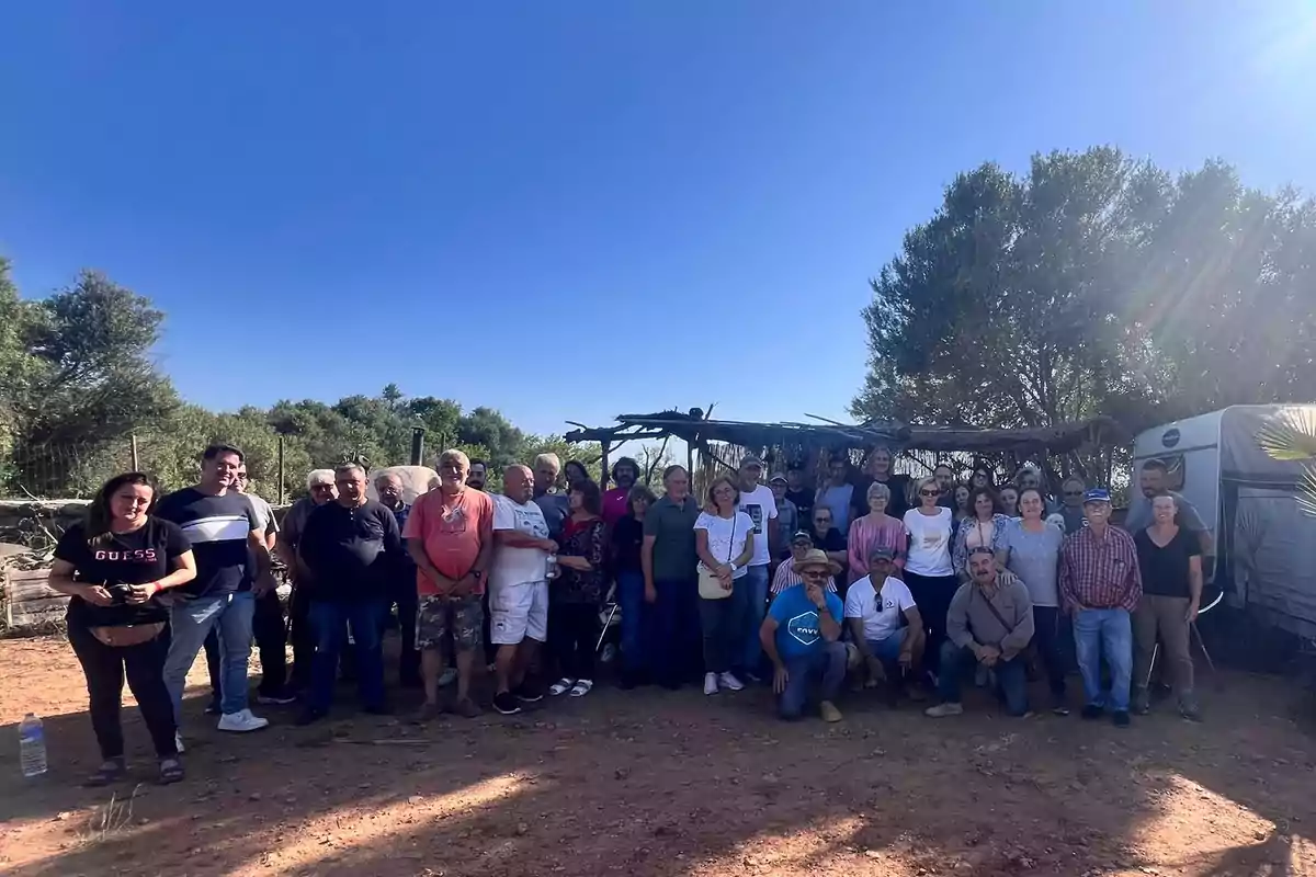Un grupo numeroso de personas posa al aire libre en un entorno rural con árboles y una estructura rústica al fondo bajo un cielo despejado Un grupo numeroso de personas posa al aire libre en un entorno rural con árboles y una estructura rústica al fondo bajo un cielo despejado