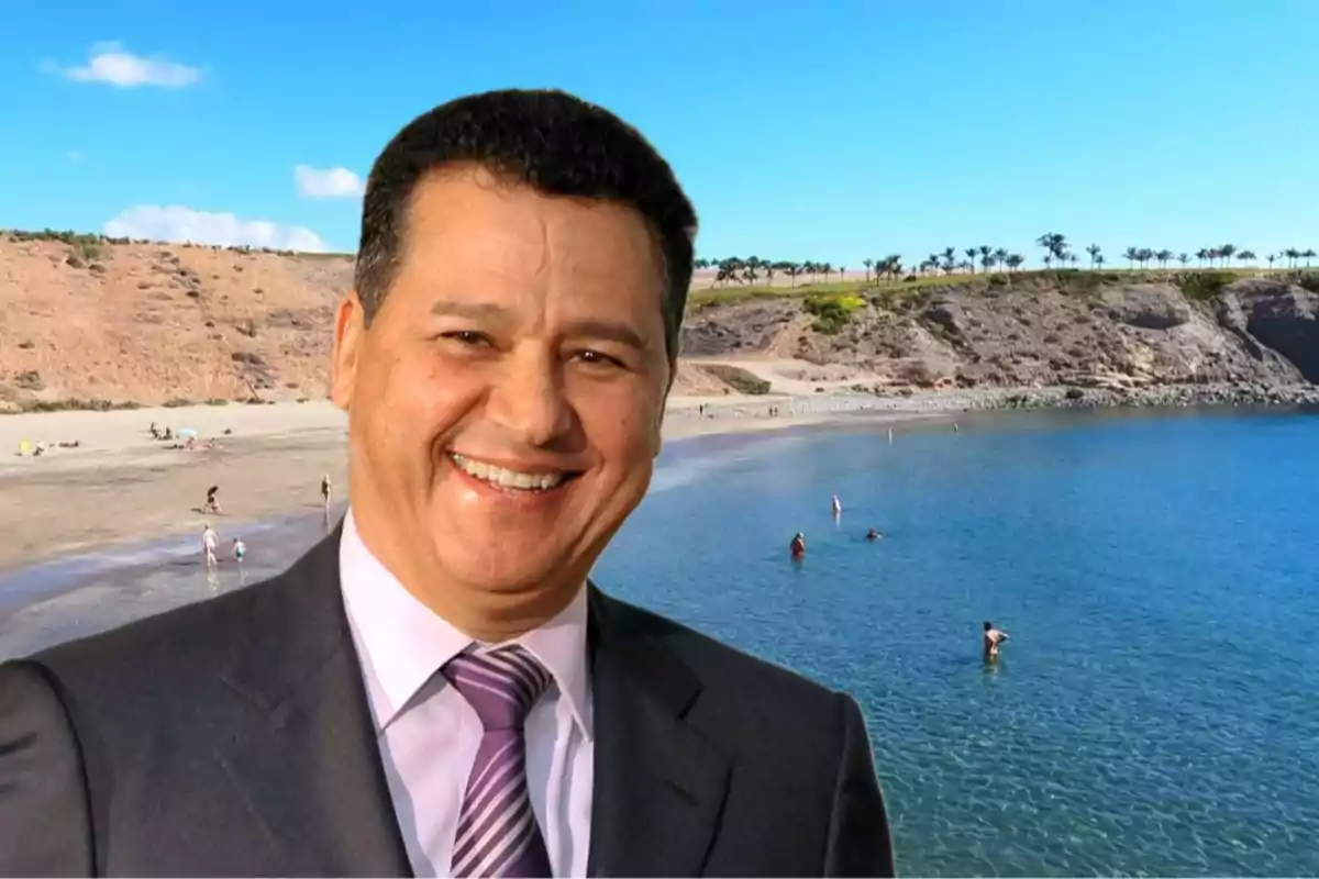Hombre sonriente con traje y corbata frente a una playa con personas disfrutando del agua y el paisaje.