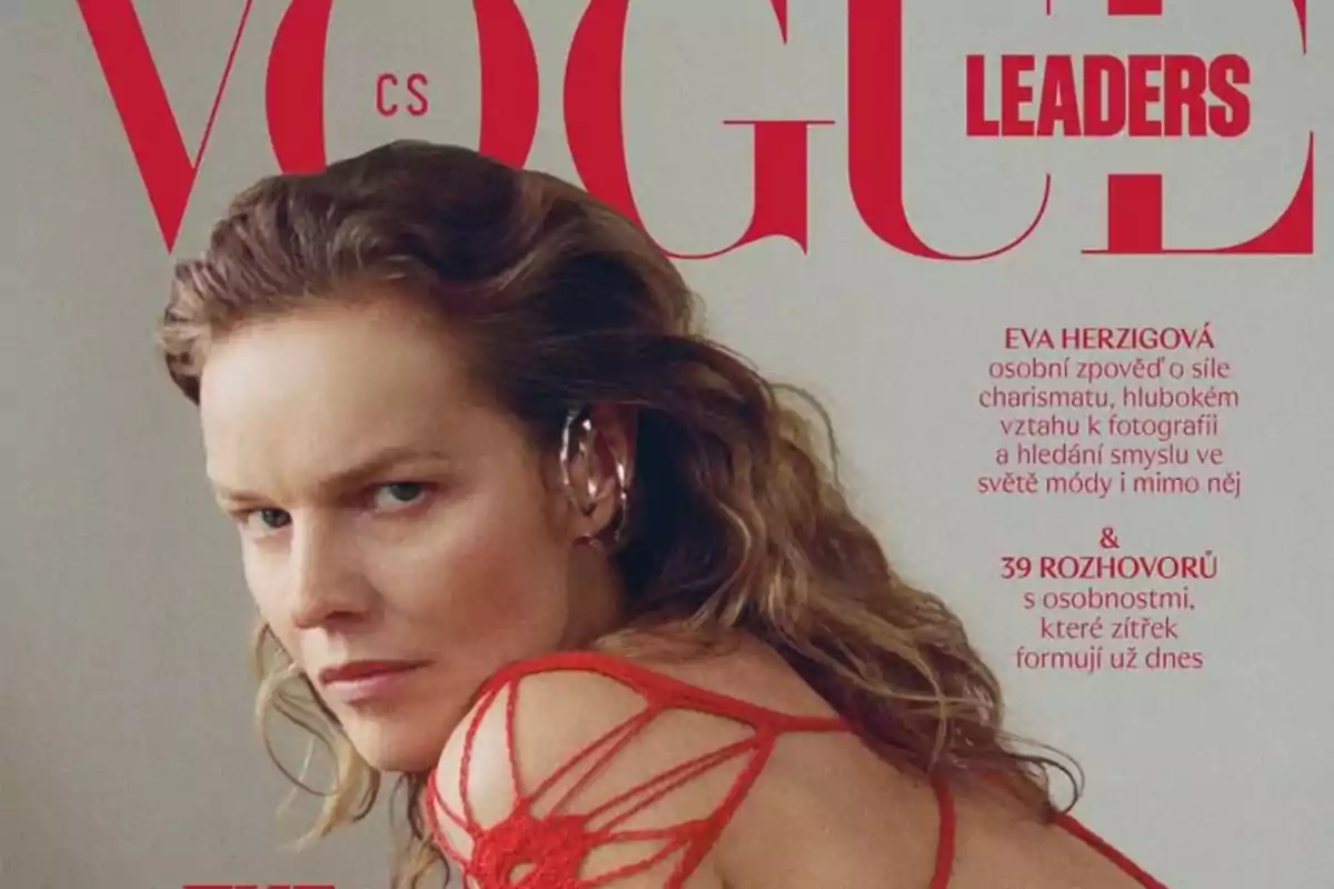 Portada de revista Vogue con una mujer de cabello ondulado rubio, usando un vestido rojo con tiras y un pendiente llamativo en la oreja, mirando de manera seria hacia la cámara Portada de revista Vogue con una mujer de cabello ondulado rubio, usando un vestido rojo con tiras y un pendiente llamativo en la oreja, mirando de manera seria hacia la cámara