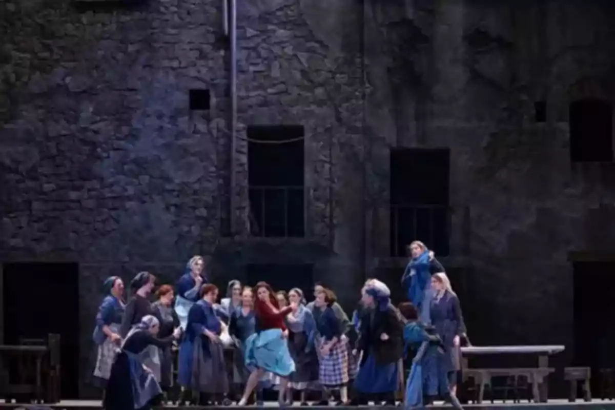 Un grupo de mujeres vestidas con ropa de época se reúne en el escenario de un teatro con un fondo de pared de piedra oscura
