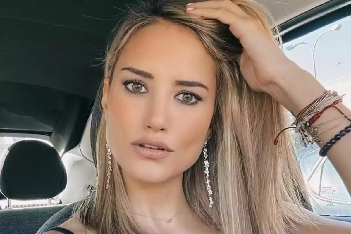 Mujer joven de cabello rubio y liso sentada en un automóvil mirando a la cámara mientras se toca el cabello y lleva varios brazaletes en la muñeca izquierda