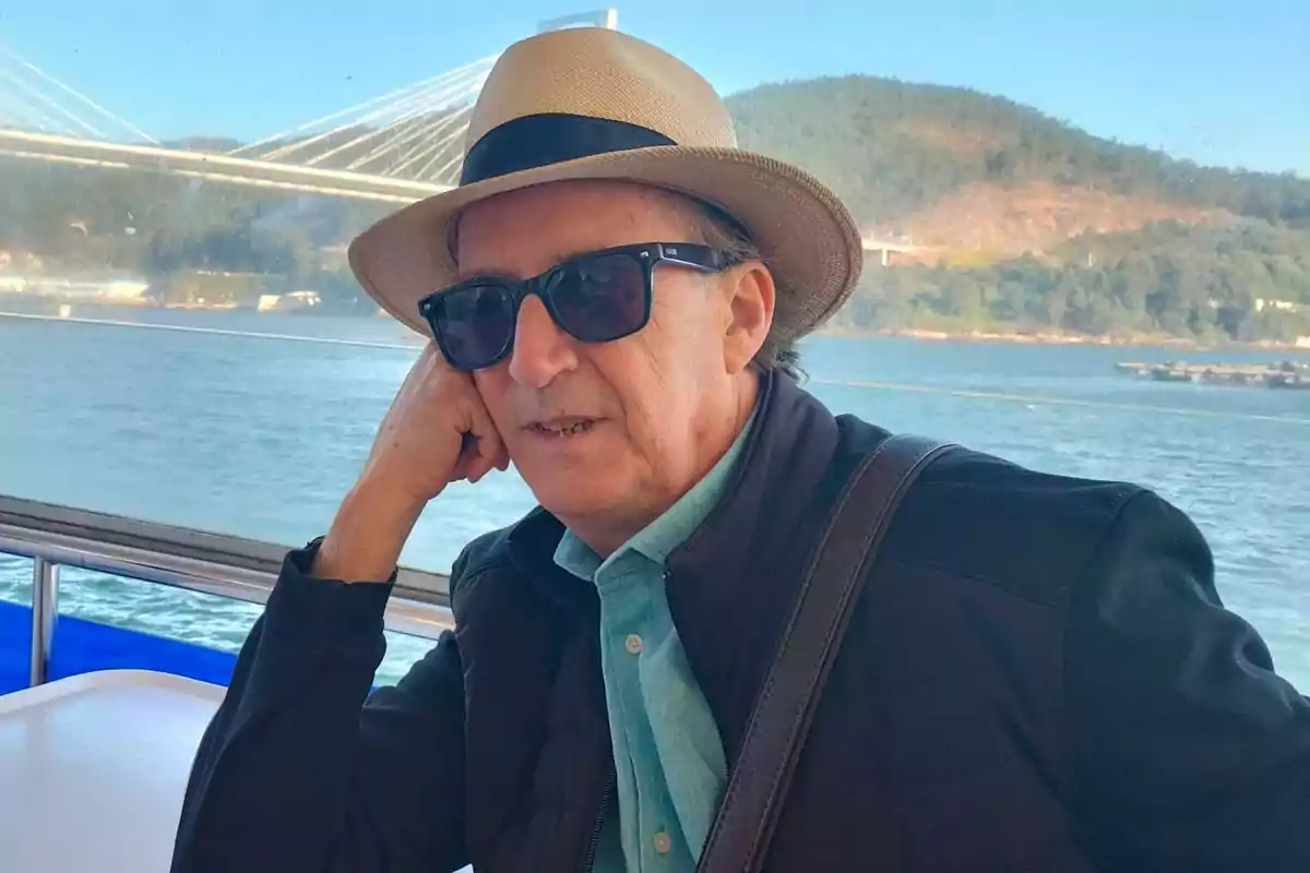 Hombre con sombrero y gafas de sol sentado junto a una ventana con vista a un río y un puente al fondo Hombre con sombrero y gafas de sol sentado junto a una ventana con vista a un río y un puente al fondo