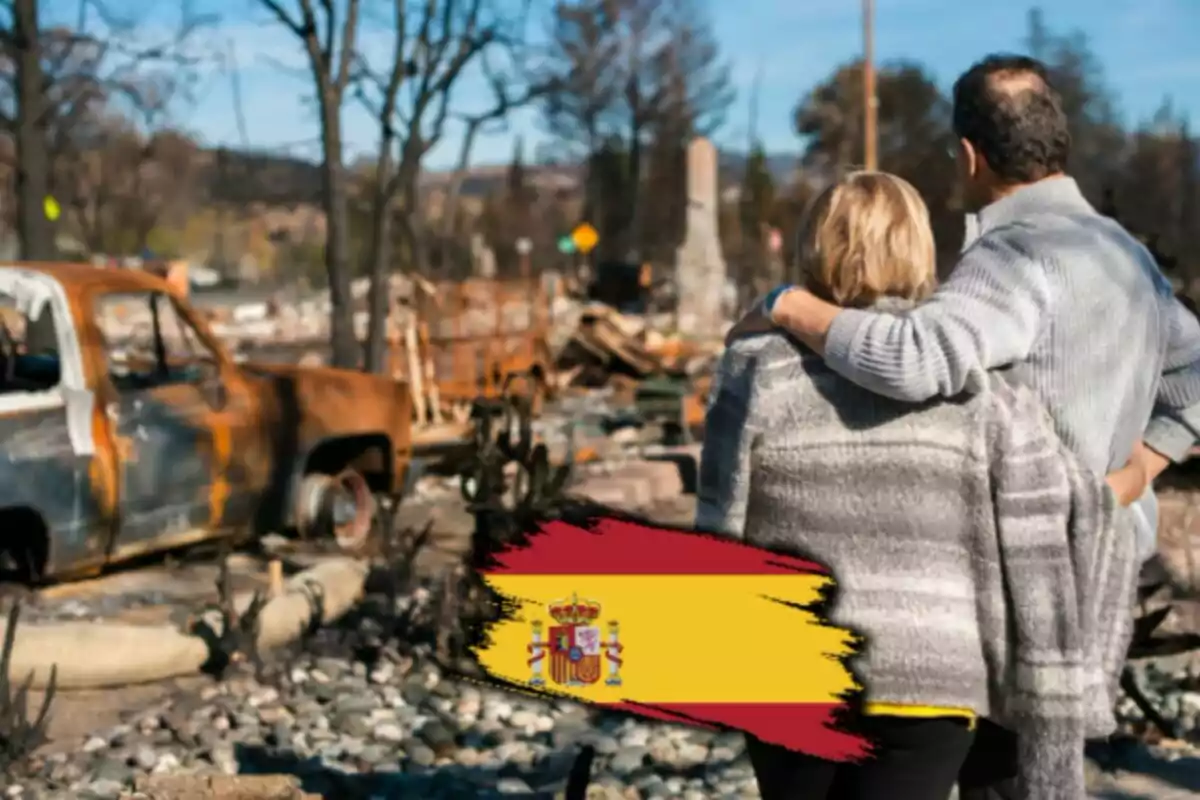 Una pareja observa un paisaje devastado con un coche quemado y árboles sin hojas mientras una bandera de España aparece en la parte inferior de la imagen.