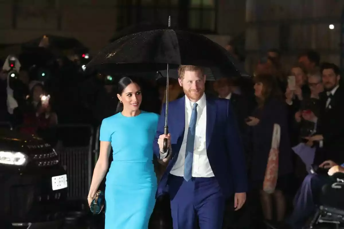 Una pareja elegante camina bajo la lluvia, compartiendo un paraguas negro mientras son fotografiados por una multitud de personas. Una pareja elegante camina bajo la lluvia, compartiendo un paraguas negro mientras son fotografiados por una multitud de personas.