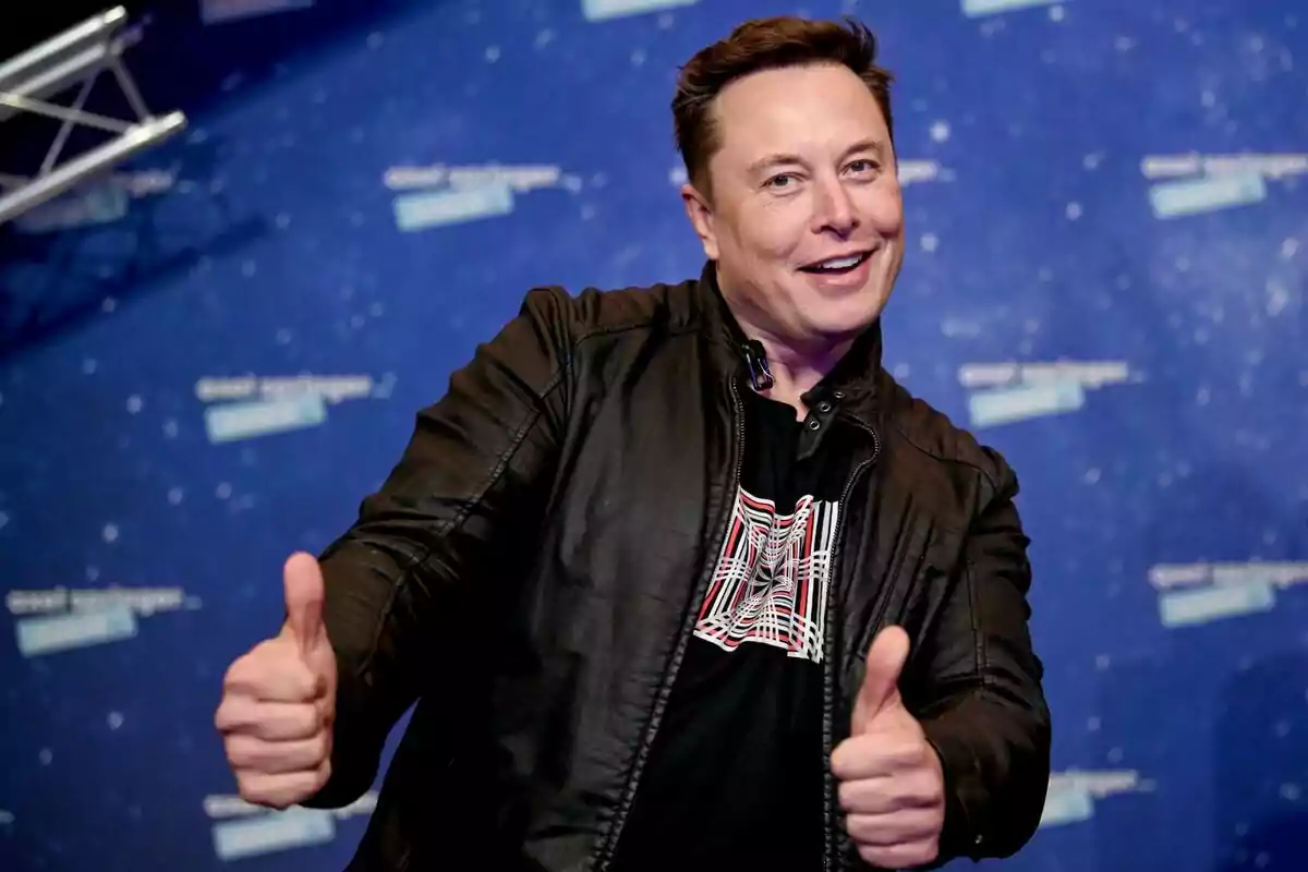A man in a black leather jacket smiles and gives both thumbs up in front of a blue background with blurred text. A man in a black leather jacket smiles and gives both thumbs up in front of a blue background with blurred text.