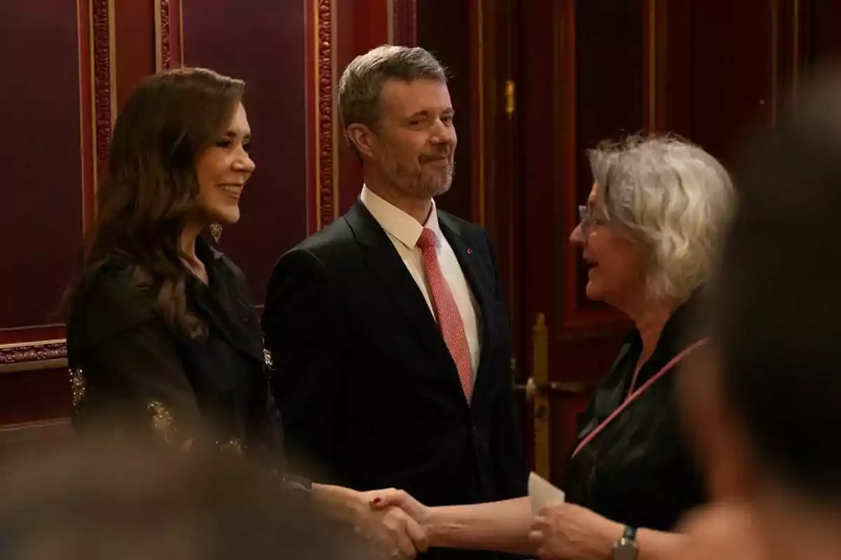 Tres personas conversan y se saludan en un ambiente formal con paredes decoradas. Tres personas conversan y se saludan en un ambiente formal con paredes decoradas.