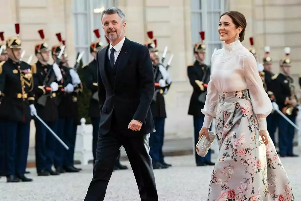 Una pareja elegantemente vestida camina frente a un grupo de guardias uniformados en un evento formal. Una pareja elegantemente vestida camina frente a un grupo de guardias uniformados en un evento formal.