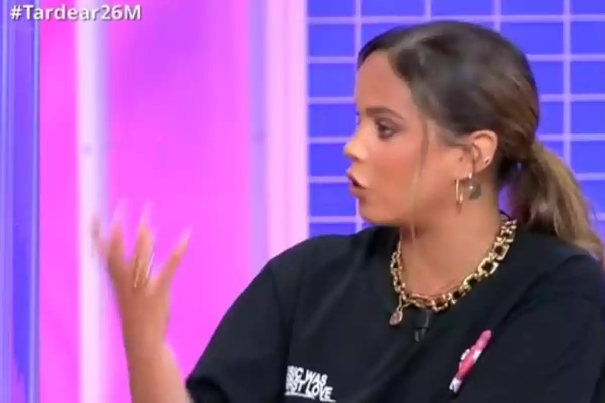 Gloria Camila joven con coleta y camiseta negra hablando y gesticulando en un set de televisión con fondo rosa y azul Gloria Camila joven con coleta y camiseta negra hablando y gesticulando en un set de televisión con fondo rosa y azul