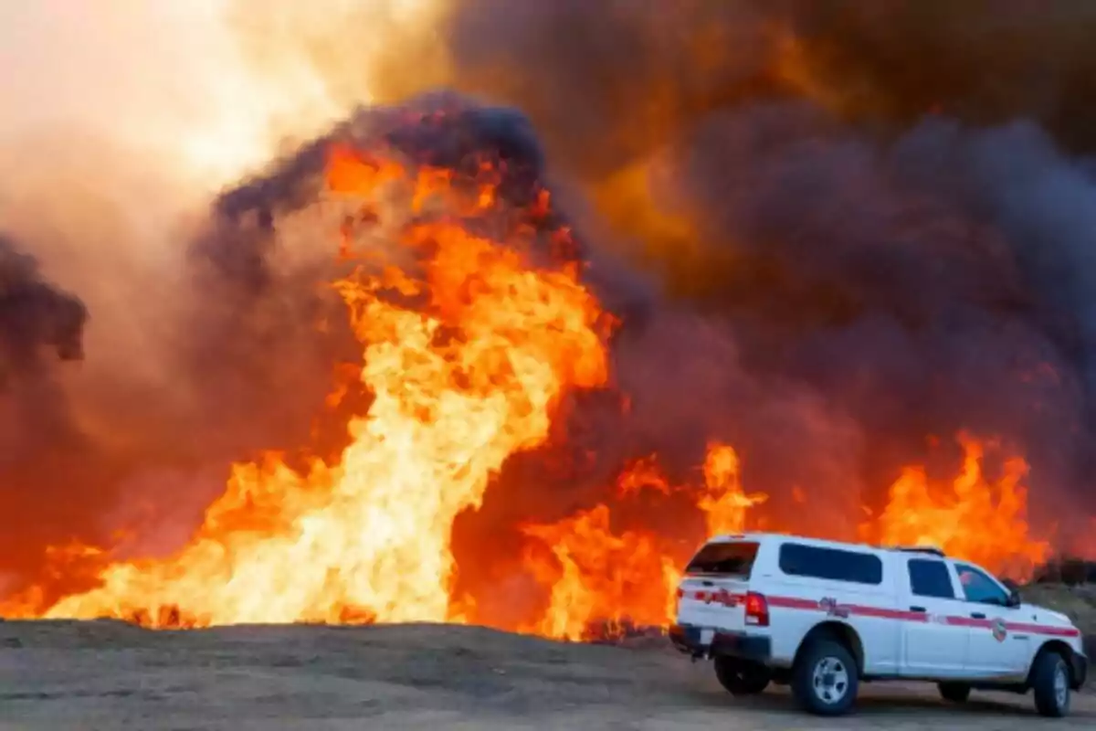 Un vehículo de emergencia estacionado cerca de un incendio forestal con grandes llamas y humo denso en el fondo en Los Ángeles. Un vehículo de emergencia estacionado cerca de un incendio forestal con grandes llamas y humo denso en el fondo en Los Ángeles.
