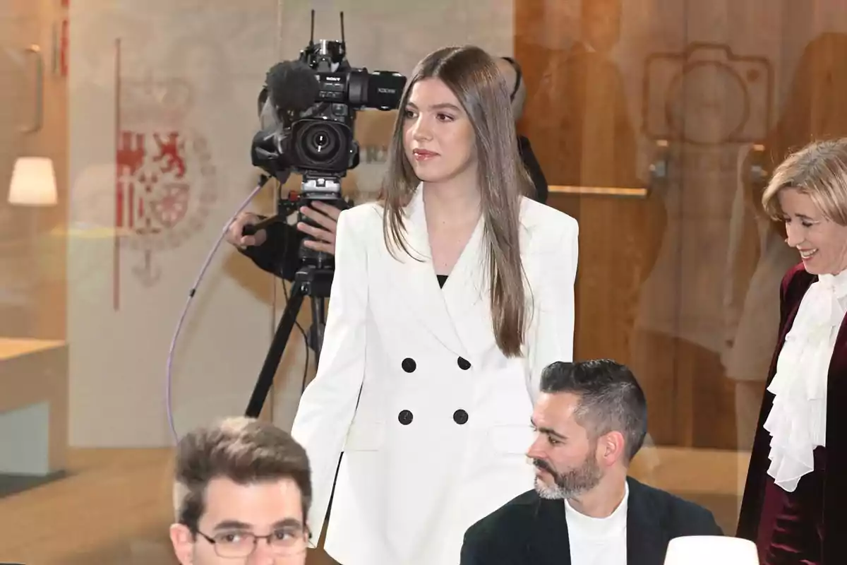 La infanta Sofía con un traje blanco está de pie frente a una cámara de televisión mientras otras personas están sentadas a su alrededor en un evento formal.